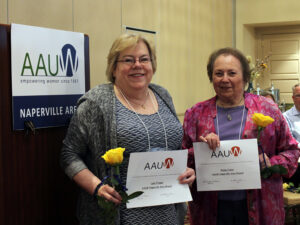 Two women pose with recognition certificates and yellow roses next to a podium with an AAUW Naperville Area sign