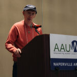 Man in orange sweater and blue baseball cap speaks from a podium labeled with an AAUW Naperville Area sign