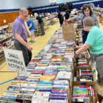 Tables full of books for sale in a school gymnasium with shoppers making selections