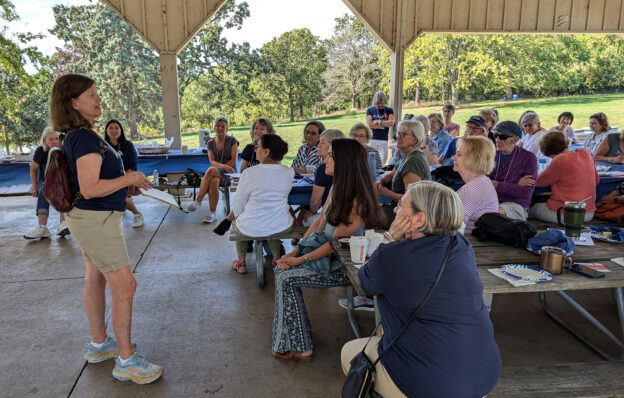 Woman speaking to group of women and men seated at picnic tables in an outdoor picnic shelter